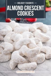 Almond crescent cookies arranged in a pile on a marble board. Powdered sugar is being dusted over the top of the cookies. Text overlay includes recipe name.