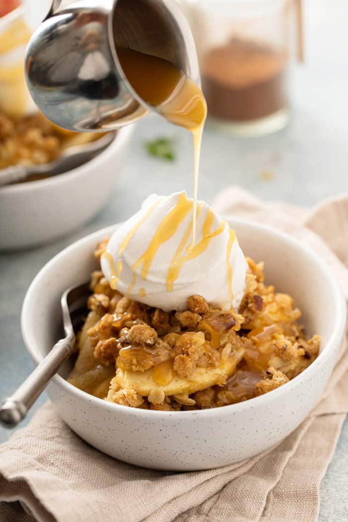 Caramel sauce being drizzled over apple pear crisp and whipped cream in a white bowl