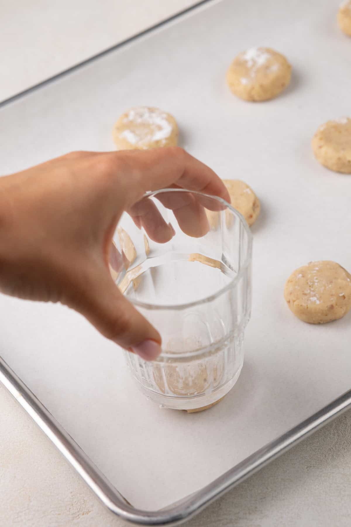 Hand using a glass to press down balls of potato chip cookie dough on a baking sheet.