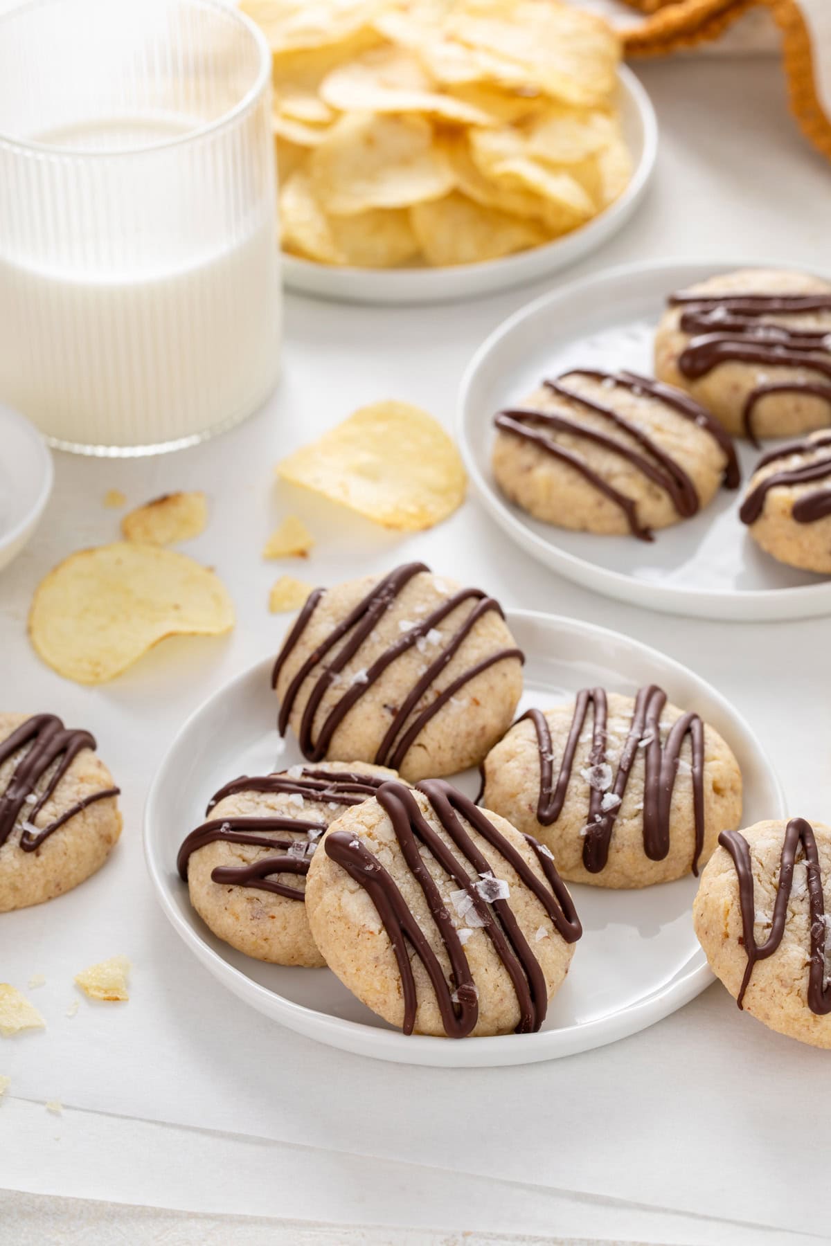 Chocolate-drizzled potato chip cookies arranged on a white plate with a glass of milk in the background.