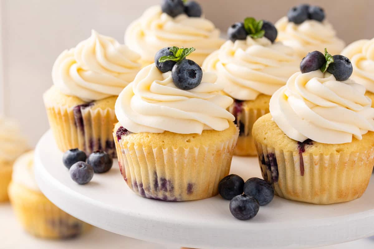 Blueberry cupcakes topped with cream cheese frosting arranged on a white cake plate.