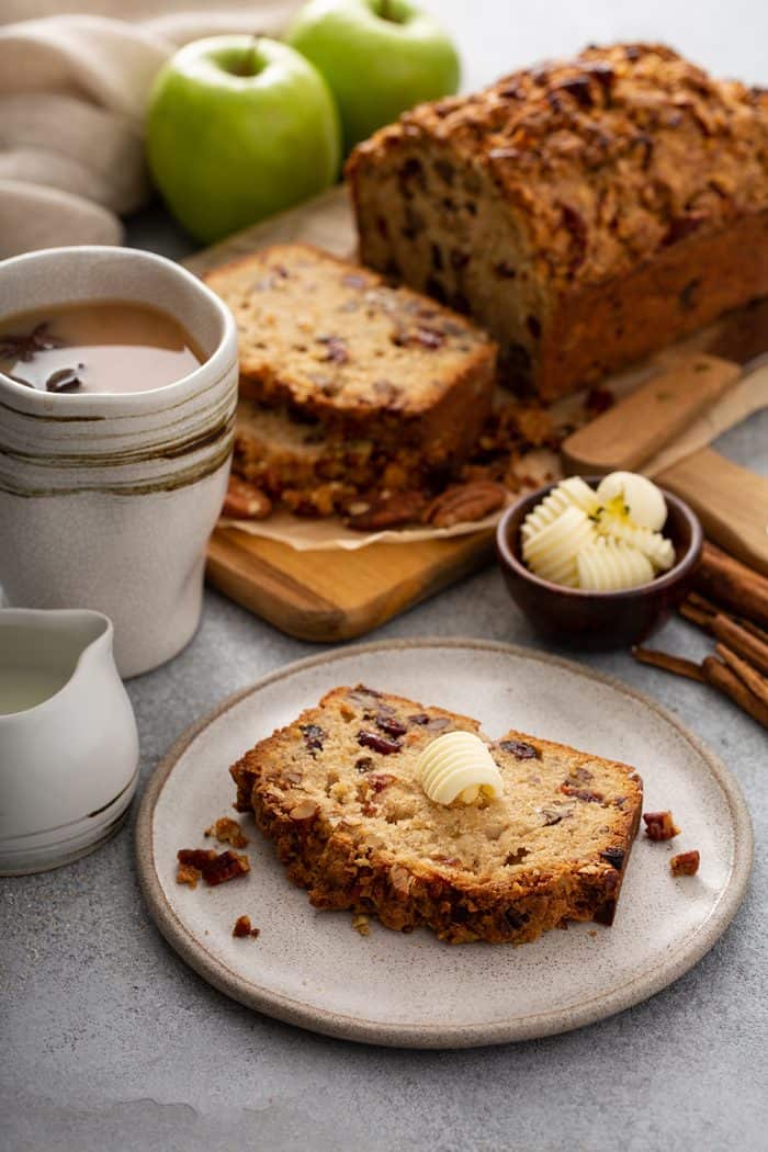 Slice of apple pie bread on a white plate with a full loaf of bread in the background