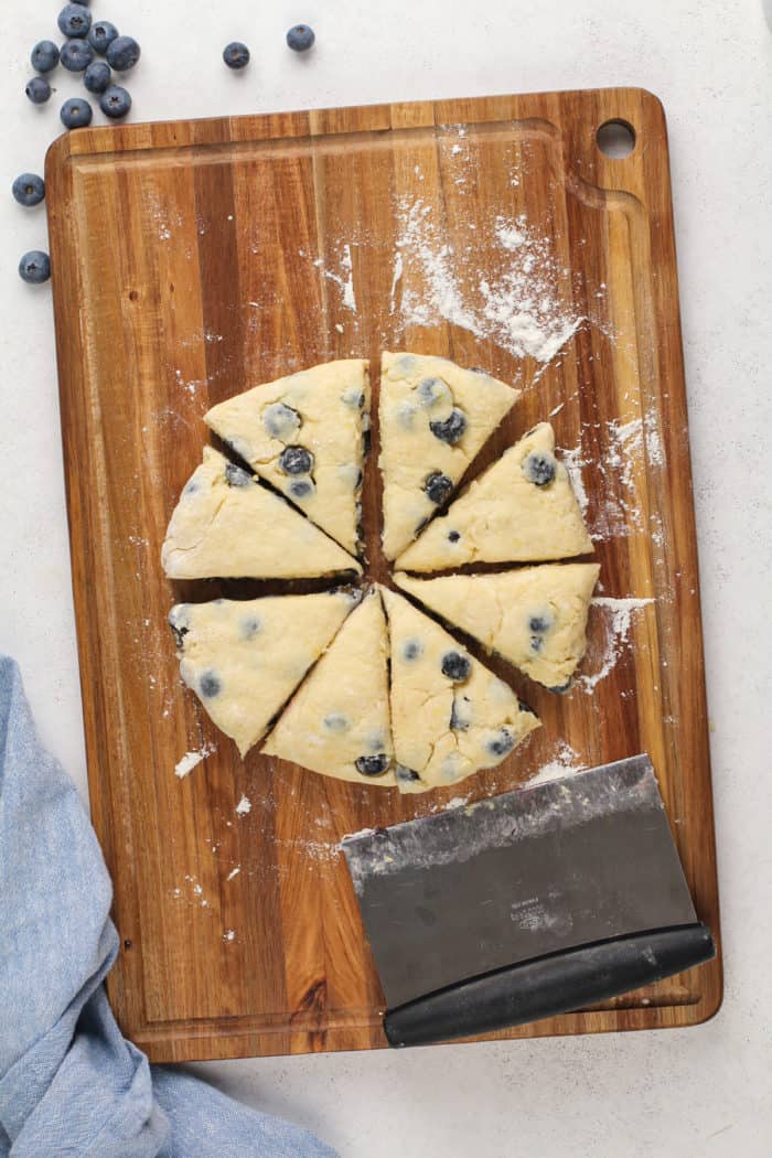 Circle of blueberry scone dough on a wooden board, cut into 8 triangles.