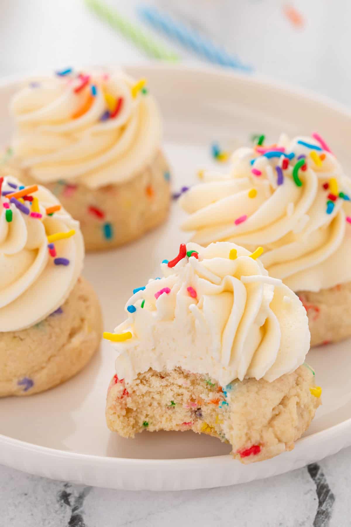 Bite taken from a frosted birthday cake cookie on a white plate, with three other cookies behind it.