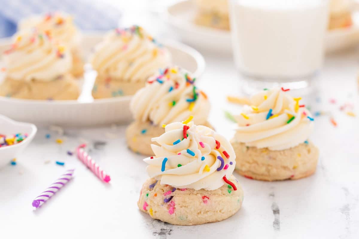 Three frosted birthday cake cookies arranged on a marble countertop with a plate of cookies in the background.