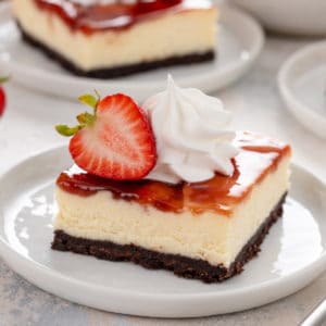 Plated strawberry cheesecake bar topped with whipped cream and a halved strawberry.