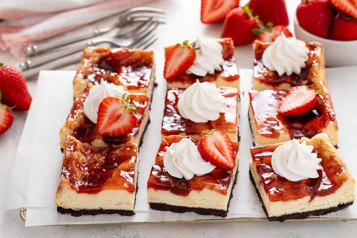 Sliced pan of strawberry cheesecake bars garnished with whipped cream and fresh strawberries.