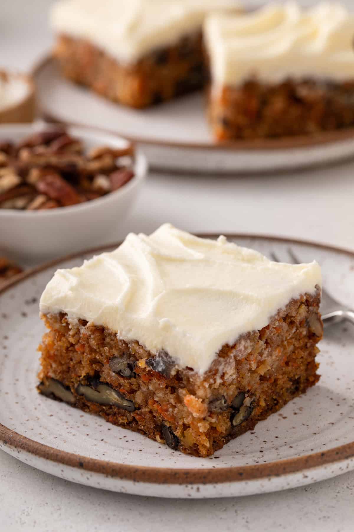 Slice of j alexander's carrot cake next to a fork on a plate, with a second plate of cake visible in the background.