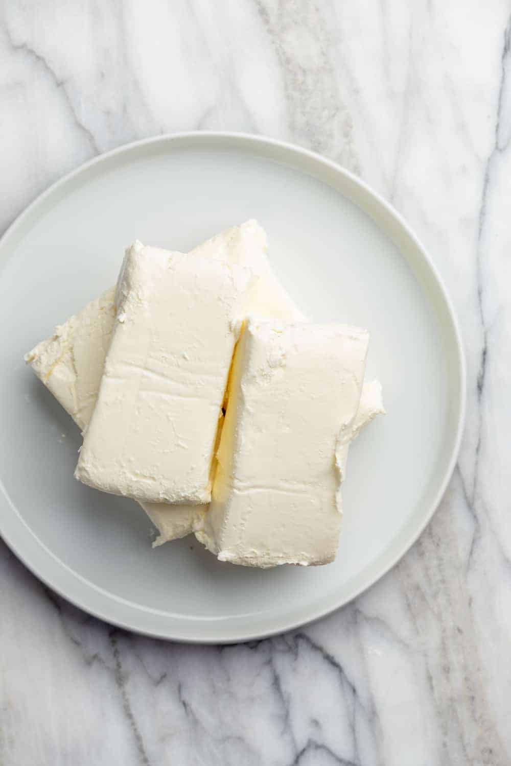Blocks of softened cream cheese on a white plate on a marble surface