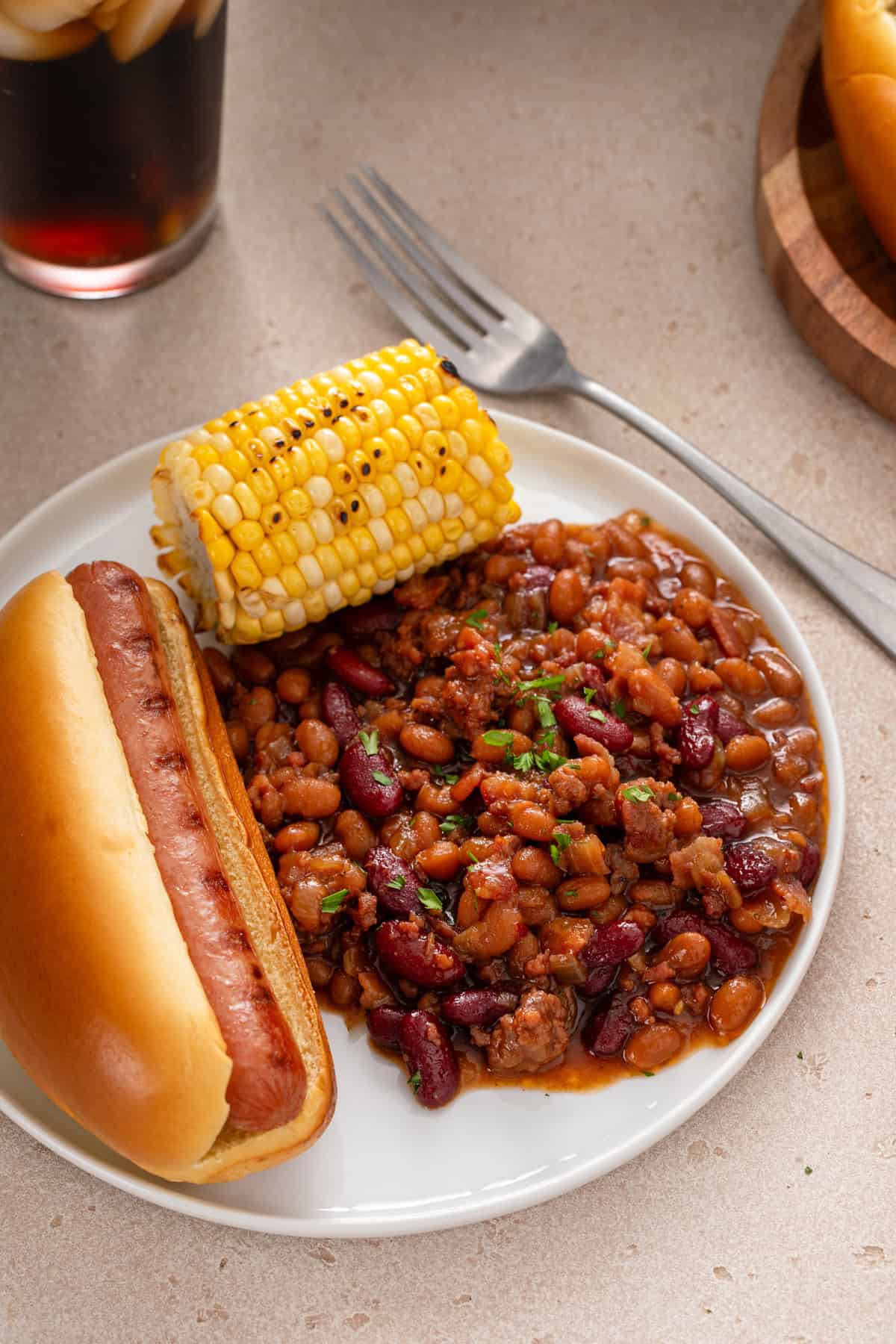 Close up image of easy baked beans on a white plate next to a hot dog and corn on the cob.