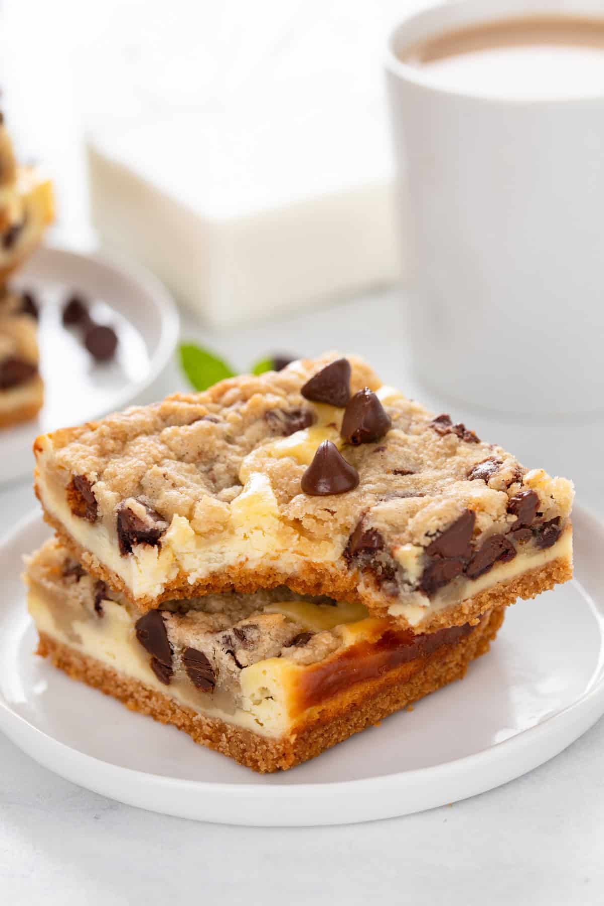 Two cookie dough cheesecake bars stacked on a white plate, with a bite taken from the corner of the top bar.