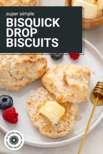 Two bisquick biscuits on a white plate next to fresh fruit. One of the biscuits is halved with butter and honey on it. Text overlay includes recipe name.
