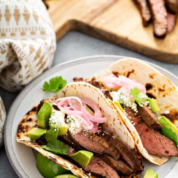 Two flank steak tacos in charred flour tortillas on a white plate with a wooden cutting board in the background
