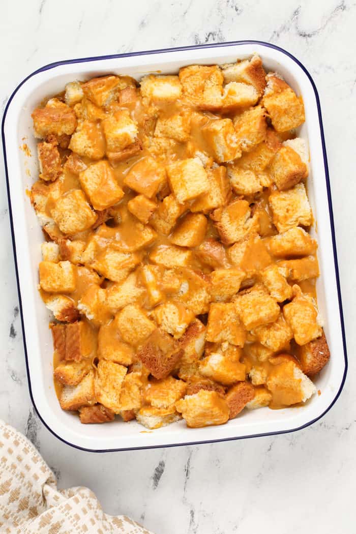 Diced challah soaked in pumpkin french toast batter in a white baking dish