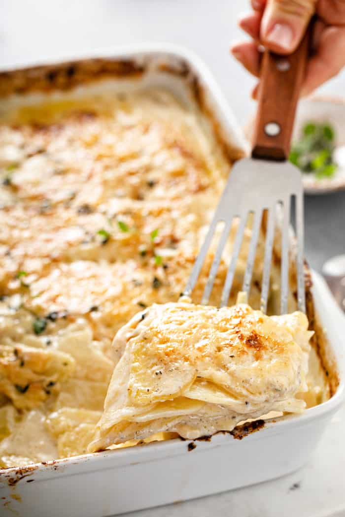Serving of potato gratin being lifted out of a white casserole dish