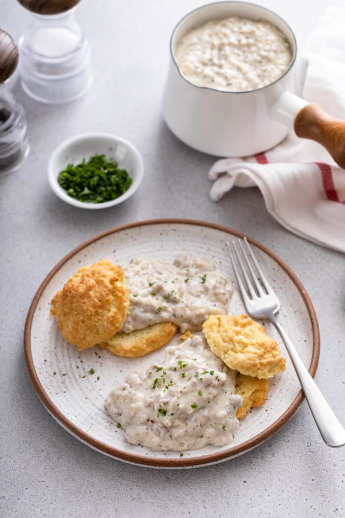 Two biscuits topped with sausage gravy on a stoneware plate. A small pot of gravy is visible in the background.