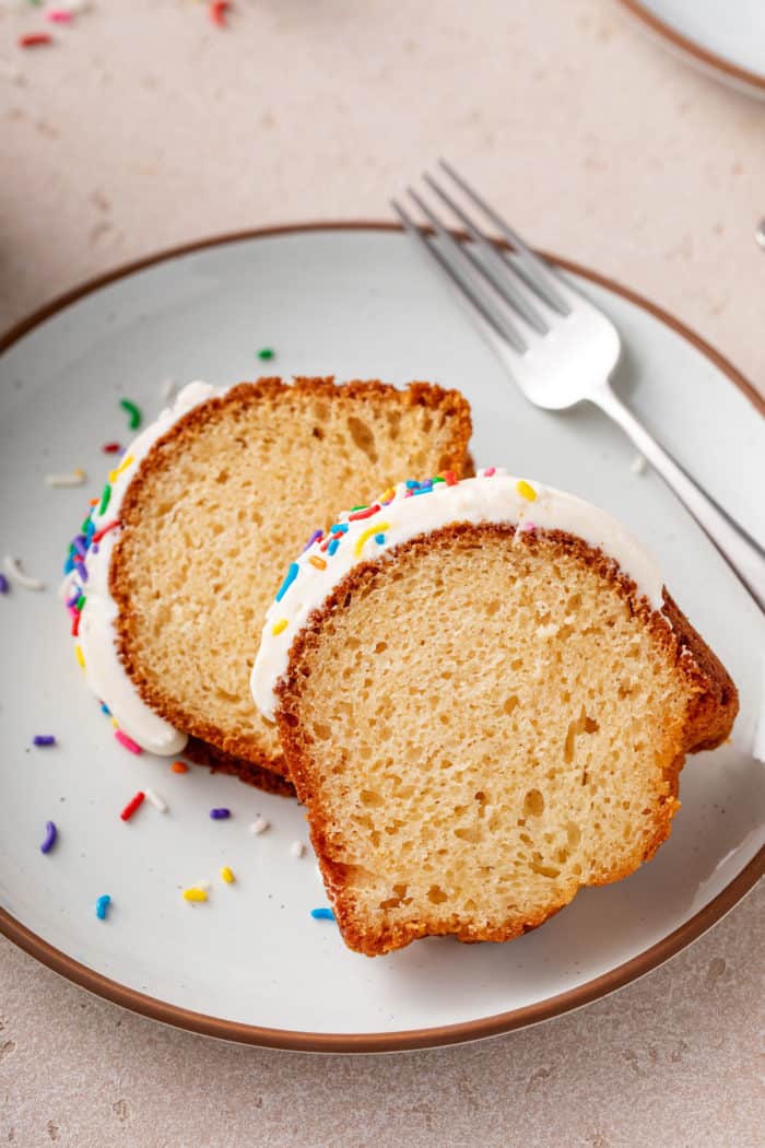 Two slices of vanilla bundt cake topped with cream cheese frosting and rainbow sprinkles laying down on a white plate.