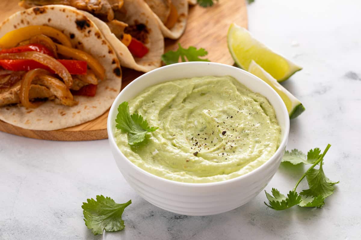 Bowl of avocado crema on a marble countertop in front of a platter of tacos.