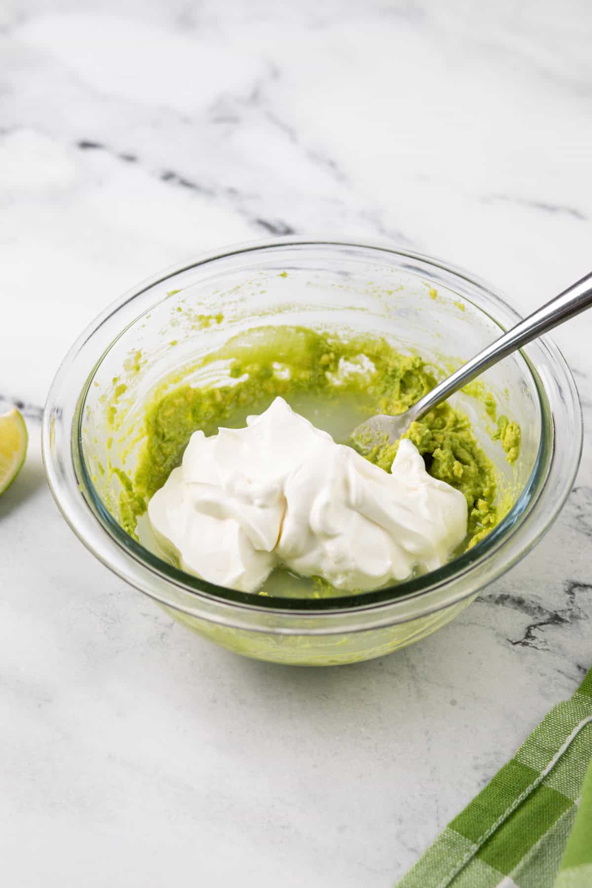 Sour cream and lime juice being added to mashed avocado in a glass bowl.