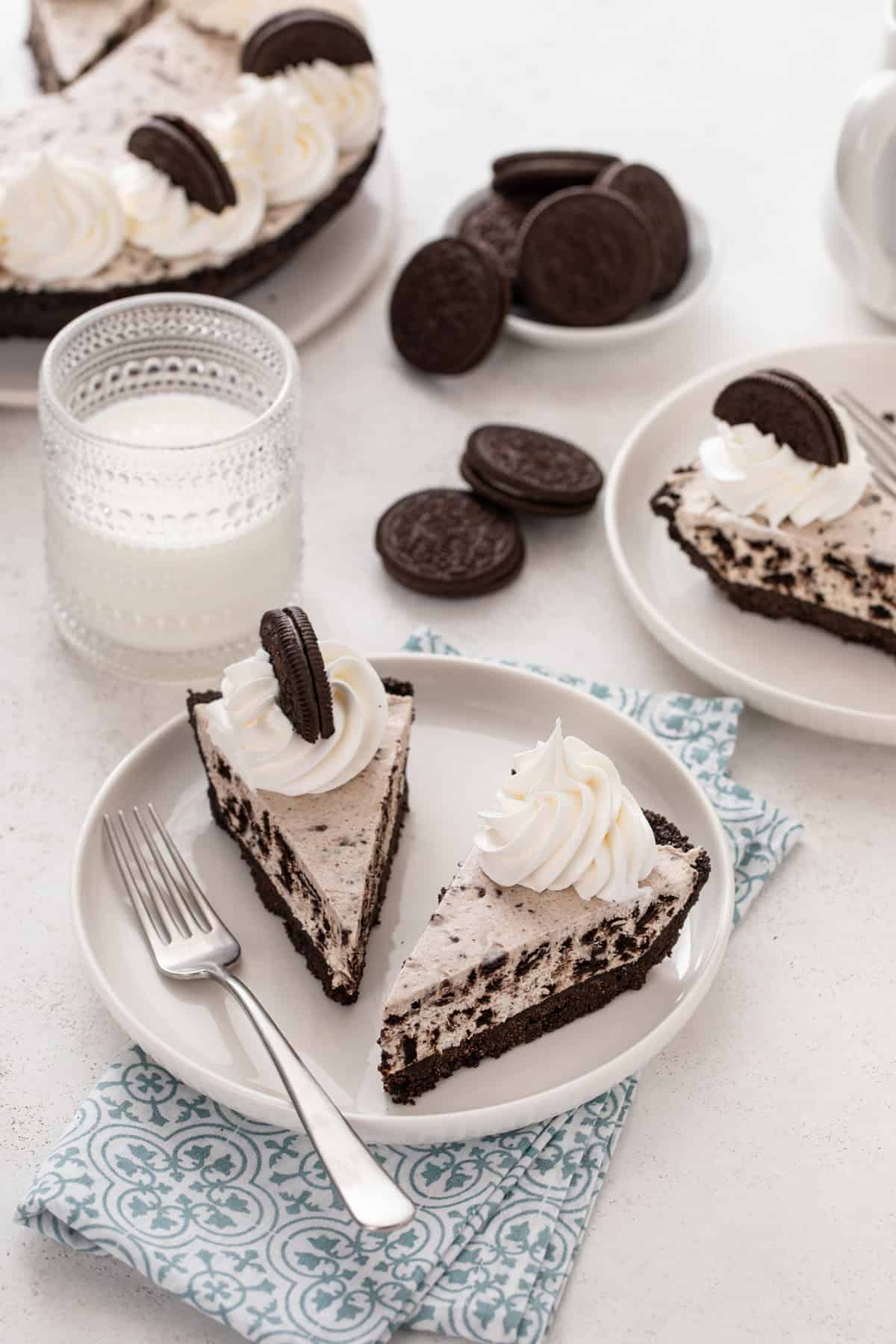 Two slices of oreo pie on a white plate with a glass of milk and more slices of pie in the background.