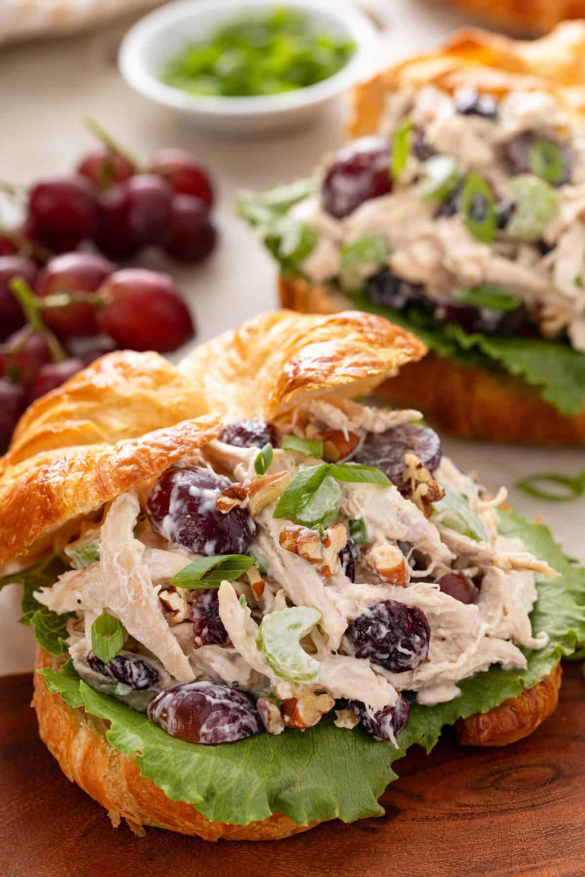 Close up image of a croissant topped with lettuce and cranberry chicken salad, with grapes and another croissant in the background.