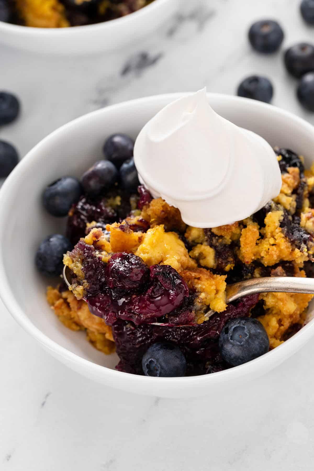 Close up of a spoon in a bowl of blueberry dump cake topped with a dollop of whipped cream.