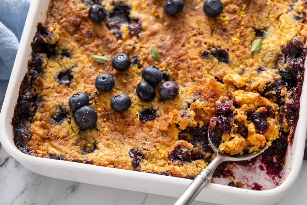 Serving spoon about to pick up blueberry dump cake from the corner of a pan.