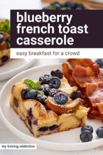 Slice of blueberry french toast casserole next to slices of bacon on a white plate. Text overlay includes recipe name.