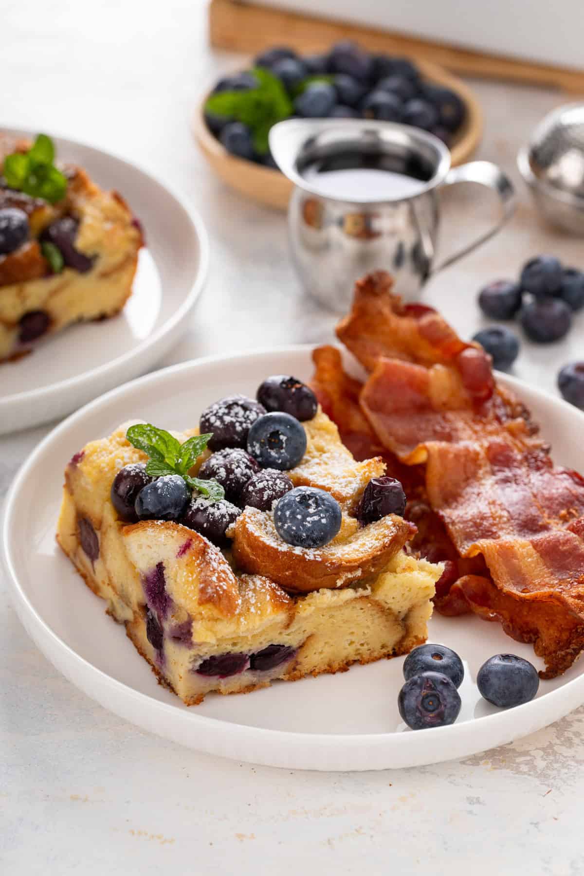 Slice of blueberry french toast casserole next to slices of bacon on a white plate.