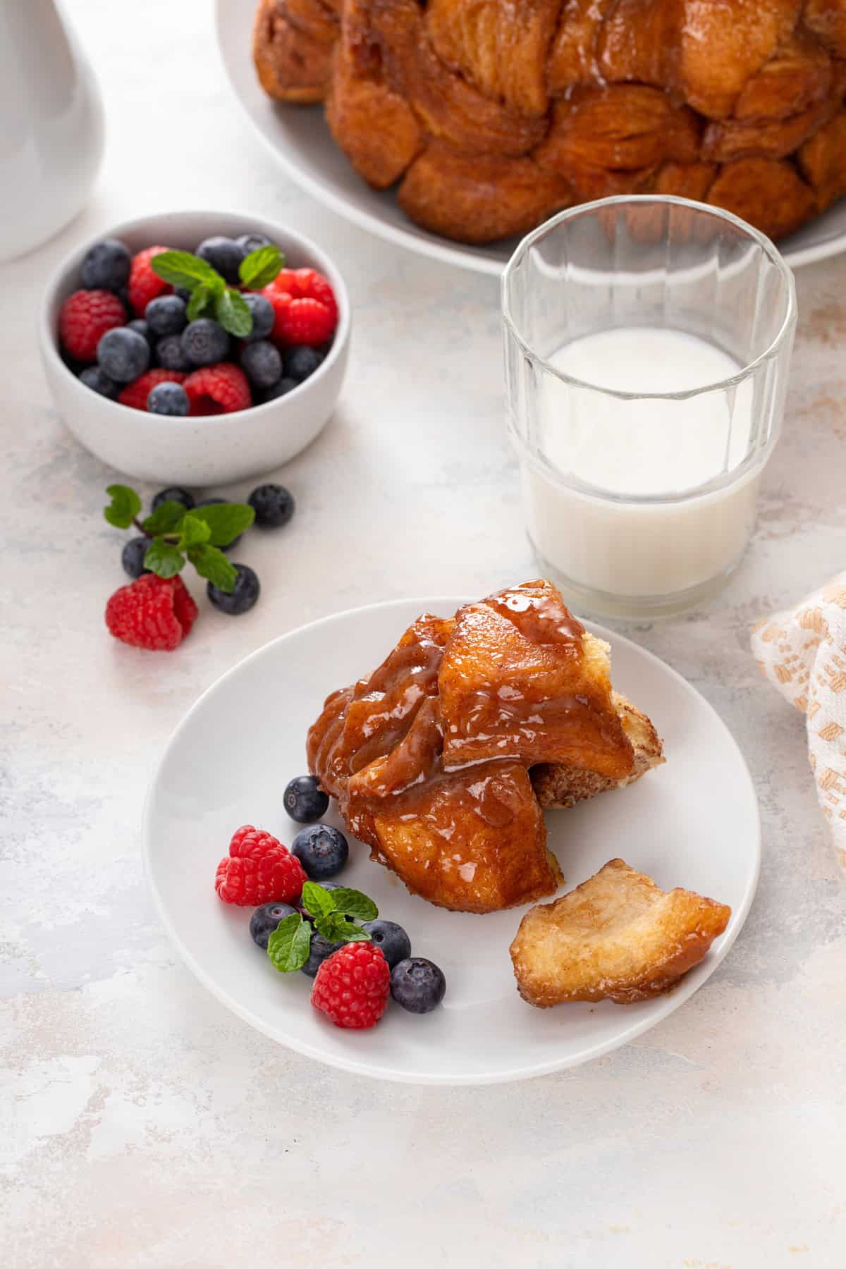 Several pieces of easy monkey bread next to fresh berries on a white plate in front of a glass of milk.
