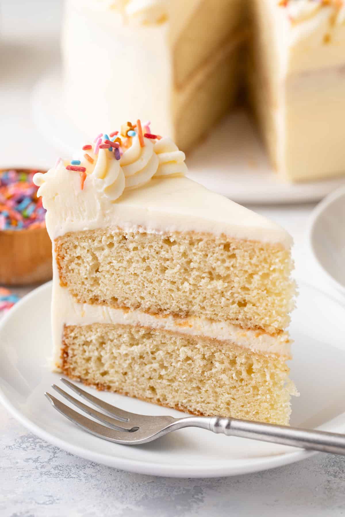 Close up of a slice of vanilla layer cake next to a fork on a white plate.