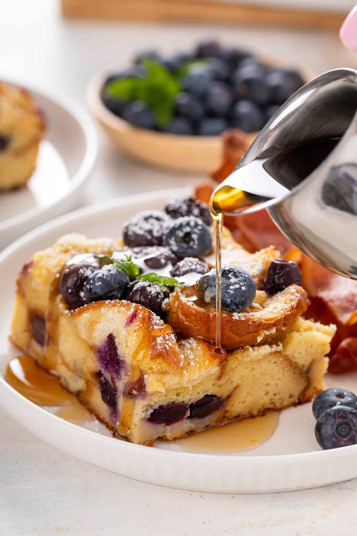 Syrup being poured over a slice of blueberry french toast casserole on a white plate.