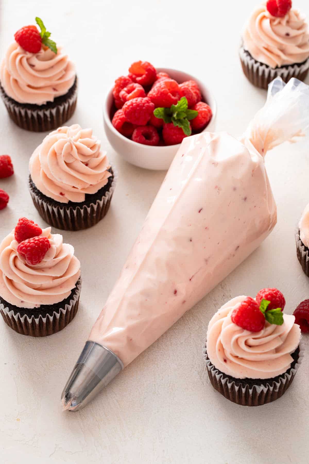 Piping bag filled with raspberry buttercream frosting surrounded by frosted chocolate cupcakes.