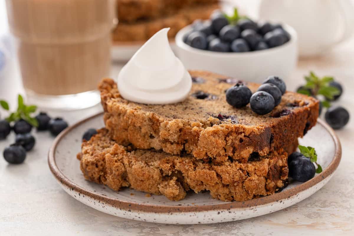 Two stacked slices of blueberry banana bread topped with a dollop of whipped cream on a plate.