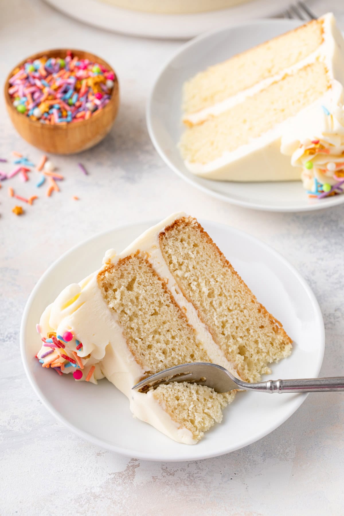 Fork cutting a corner from a slice of vanilla layer cake on a white plate.