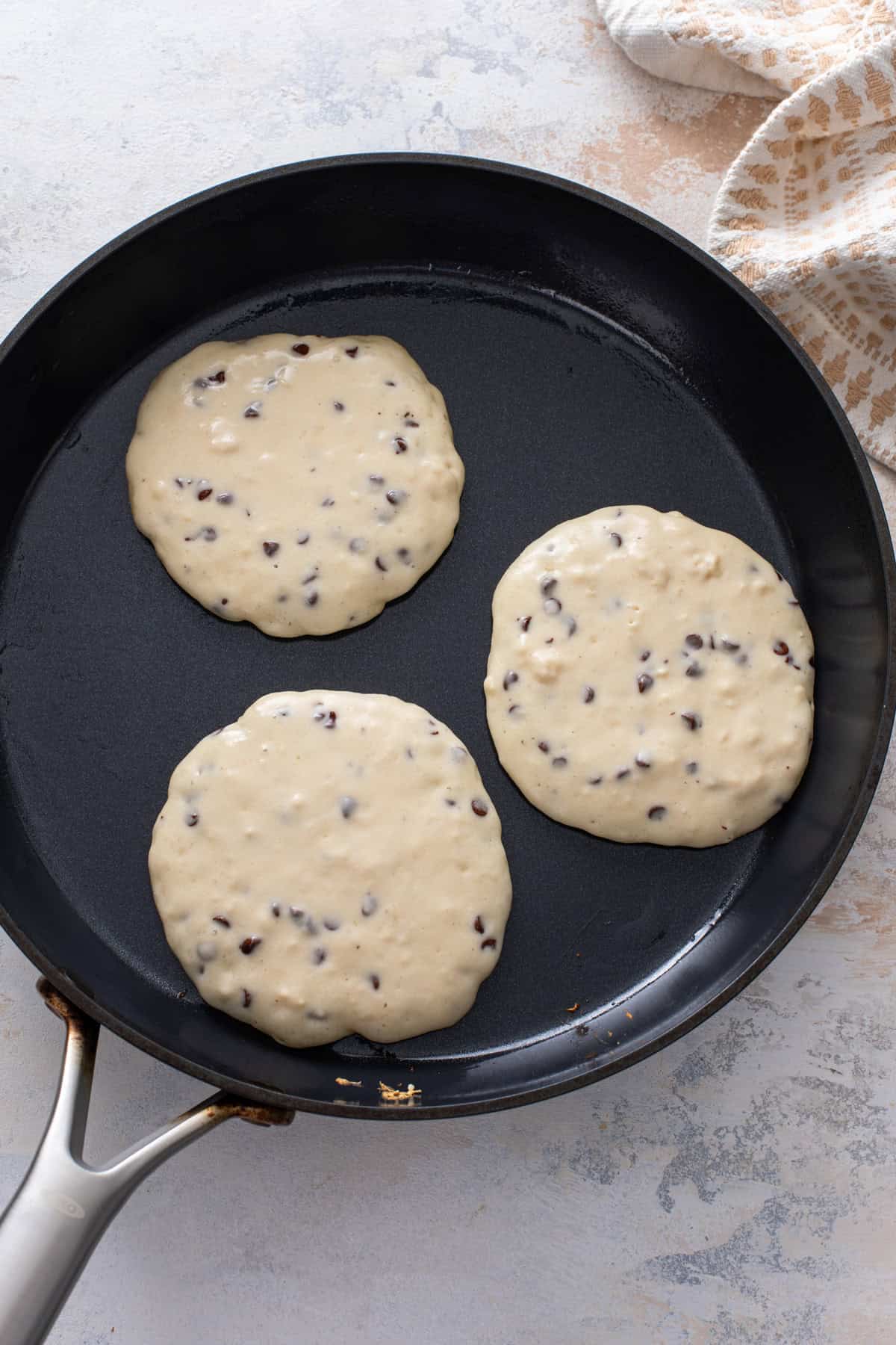 Three chocolate chip pancakes cooking in a skillet.