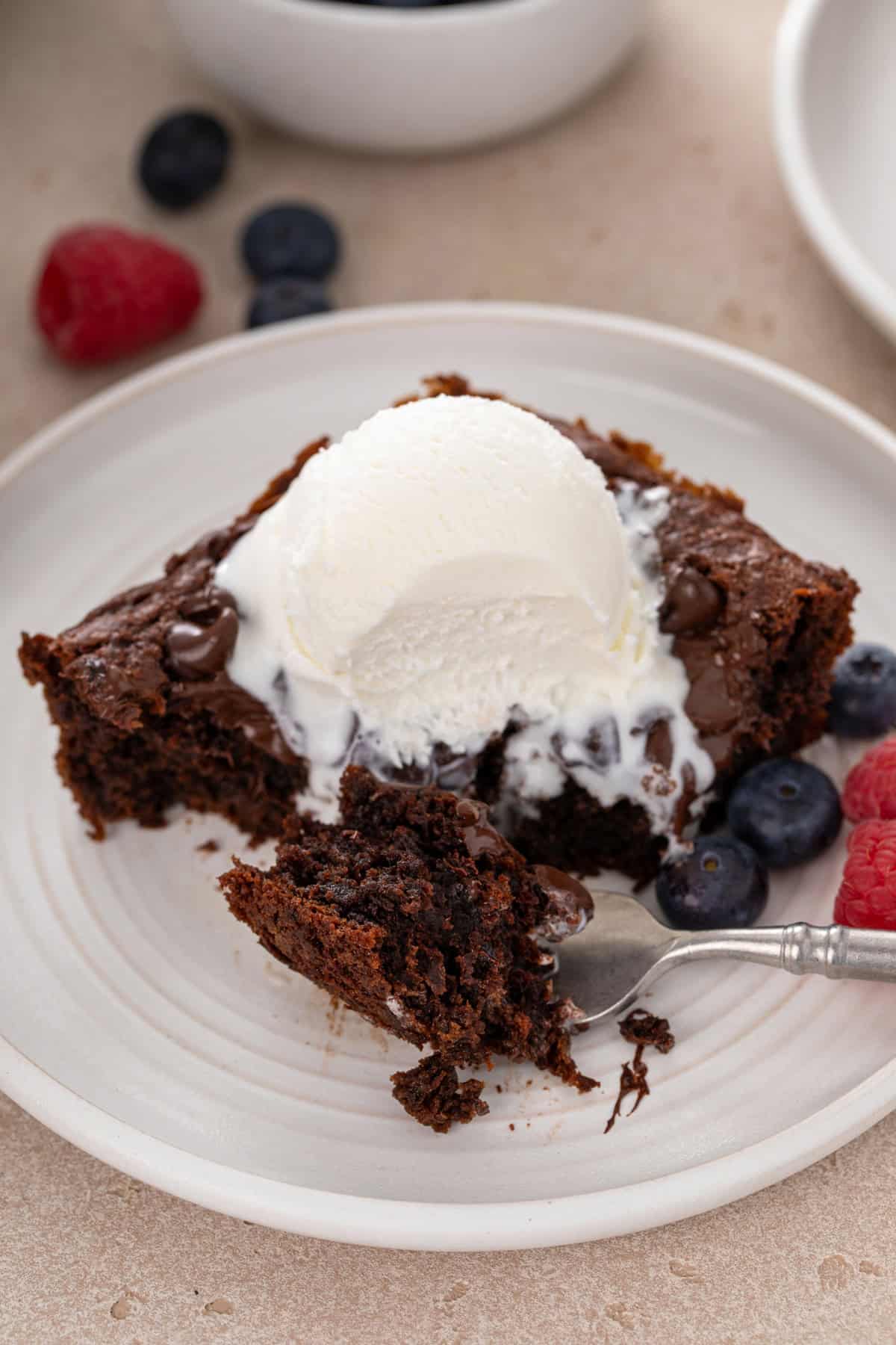Fork holding a bite of chocolate dump cake next to a slice of the cake on a white plate.