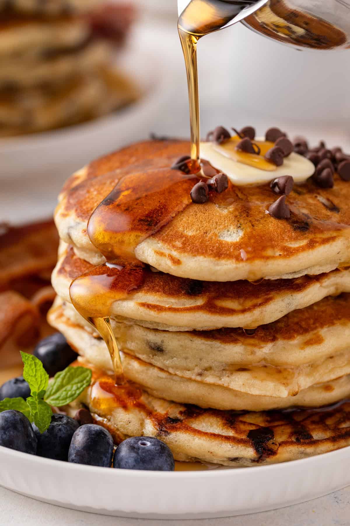 Syrup being poured over a stack of chocolate chip pancakes.