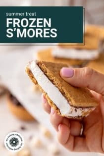 Hand holding up a frozen s'more to the camera. text overlay includes recipe name.