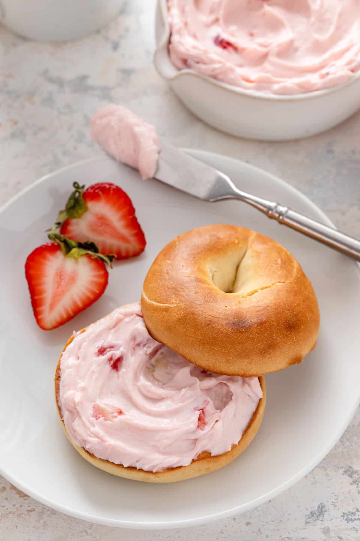 Close up of a bagel spread with strawberry cream cheese on a white plate.