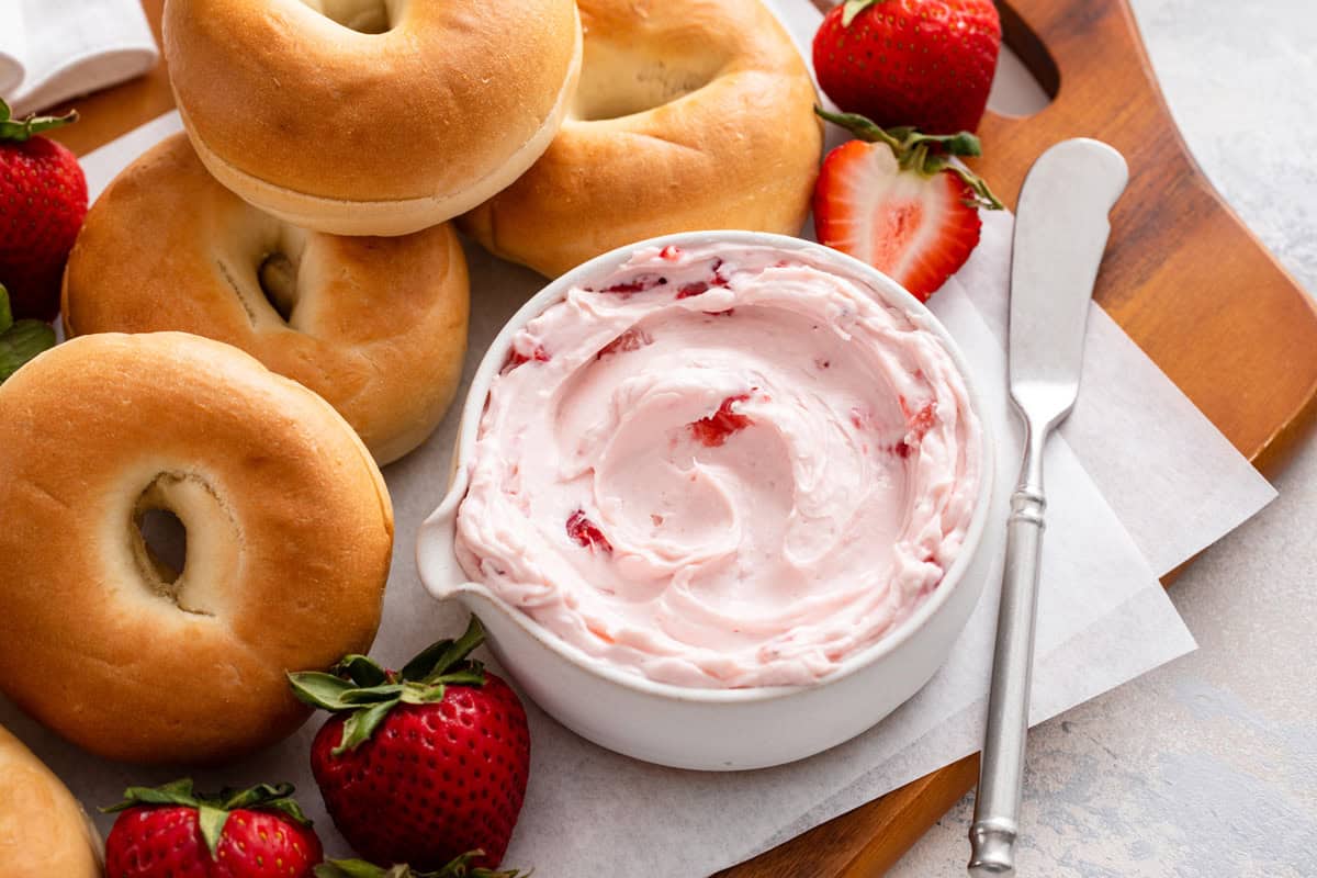 Close up of a white bowl of strawberry cream cheese surrounded by bagels.