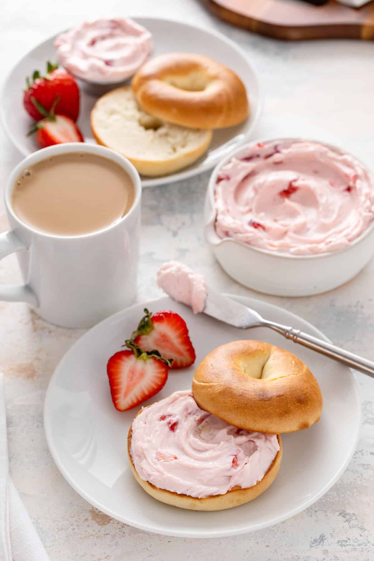 White plate filled with a toasted bagel topped with strawberry cream cheese next to a cup of coffee.