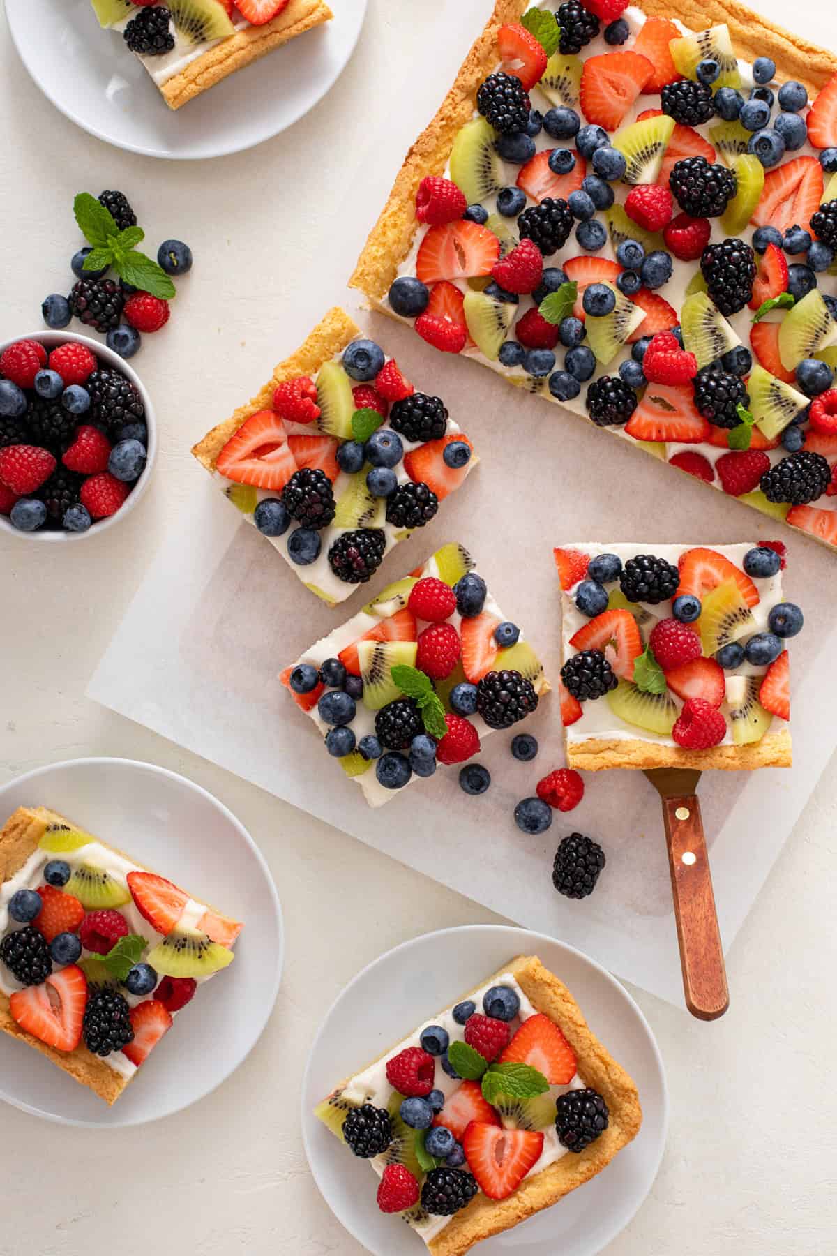 Overhead view of slices of fruit pizza, with a cake server about to lift up one of the slices.