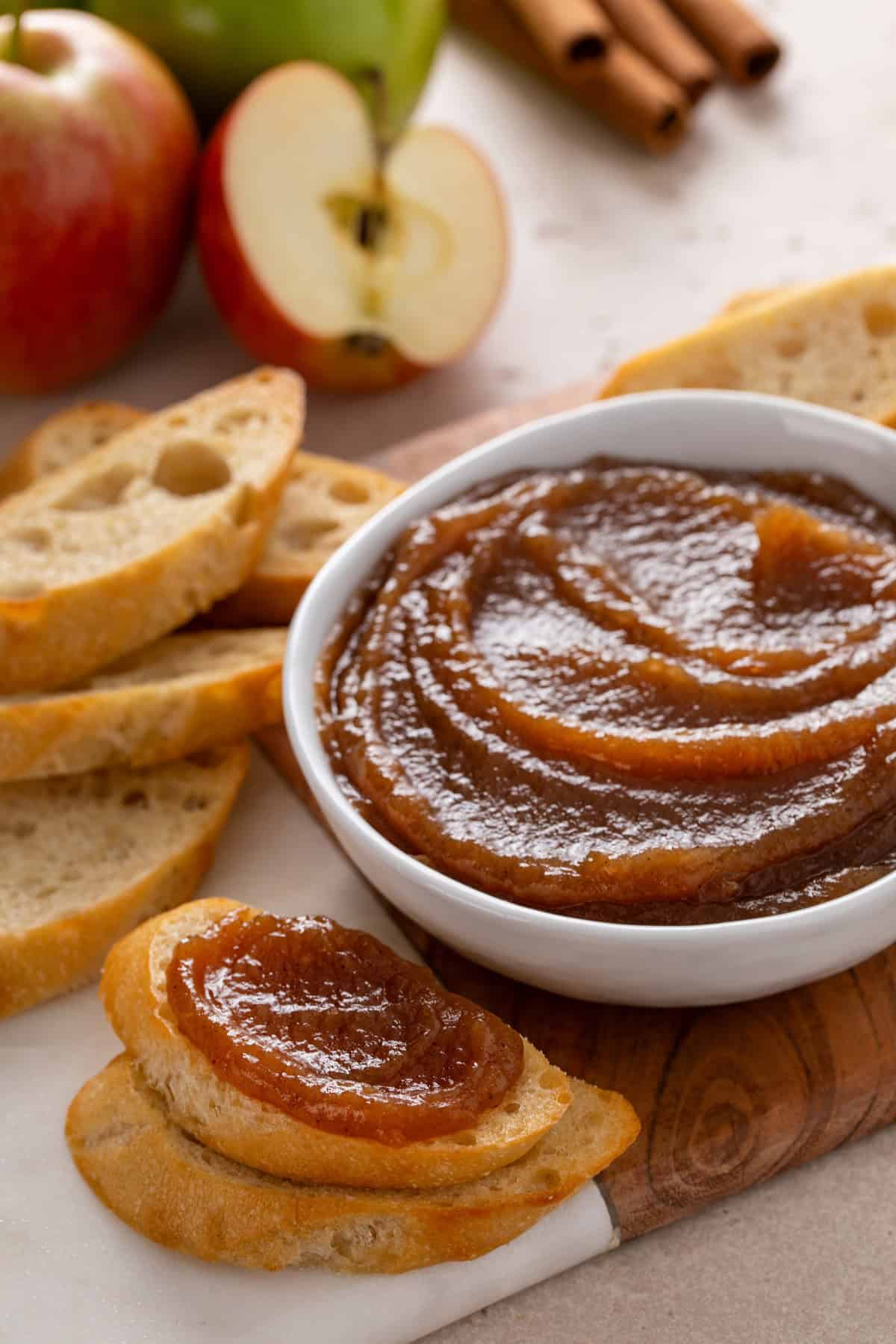 Slice of bread topped with apple butter set next to a bowl of apple butter on a wooden board.