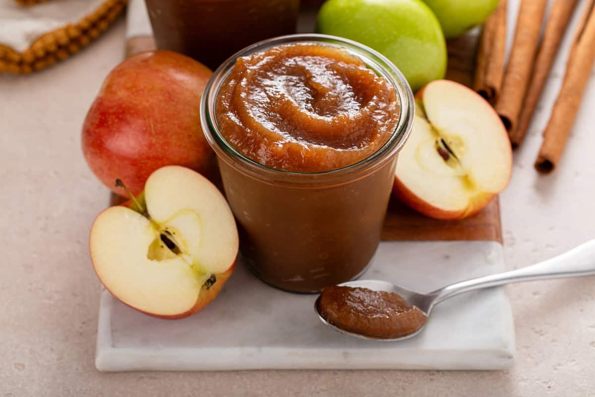 Jar of apple butter set on a marble board next to a spoonful of apple butter.