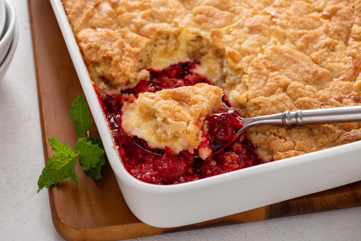 Baking dish of cherry cheesecake cake with a spoon about to serve a portion out of the corner.
