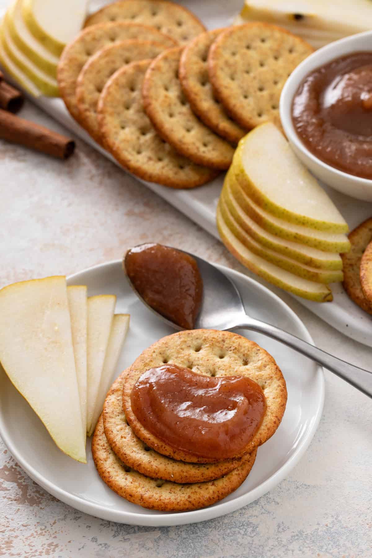 Crackers, sliced pears, and a spoon of pear butter on a white plate. One of the crackers is topped with pear butter.