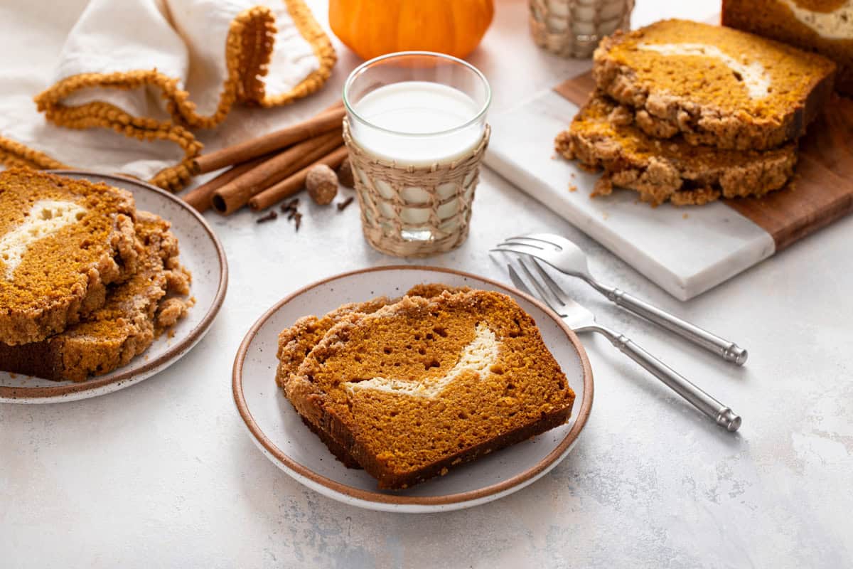 Two plates of pumpkin cream cheese bread and a glass of milk in front of a sliced loaf of the bread.