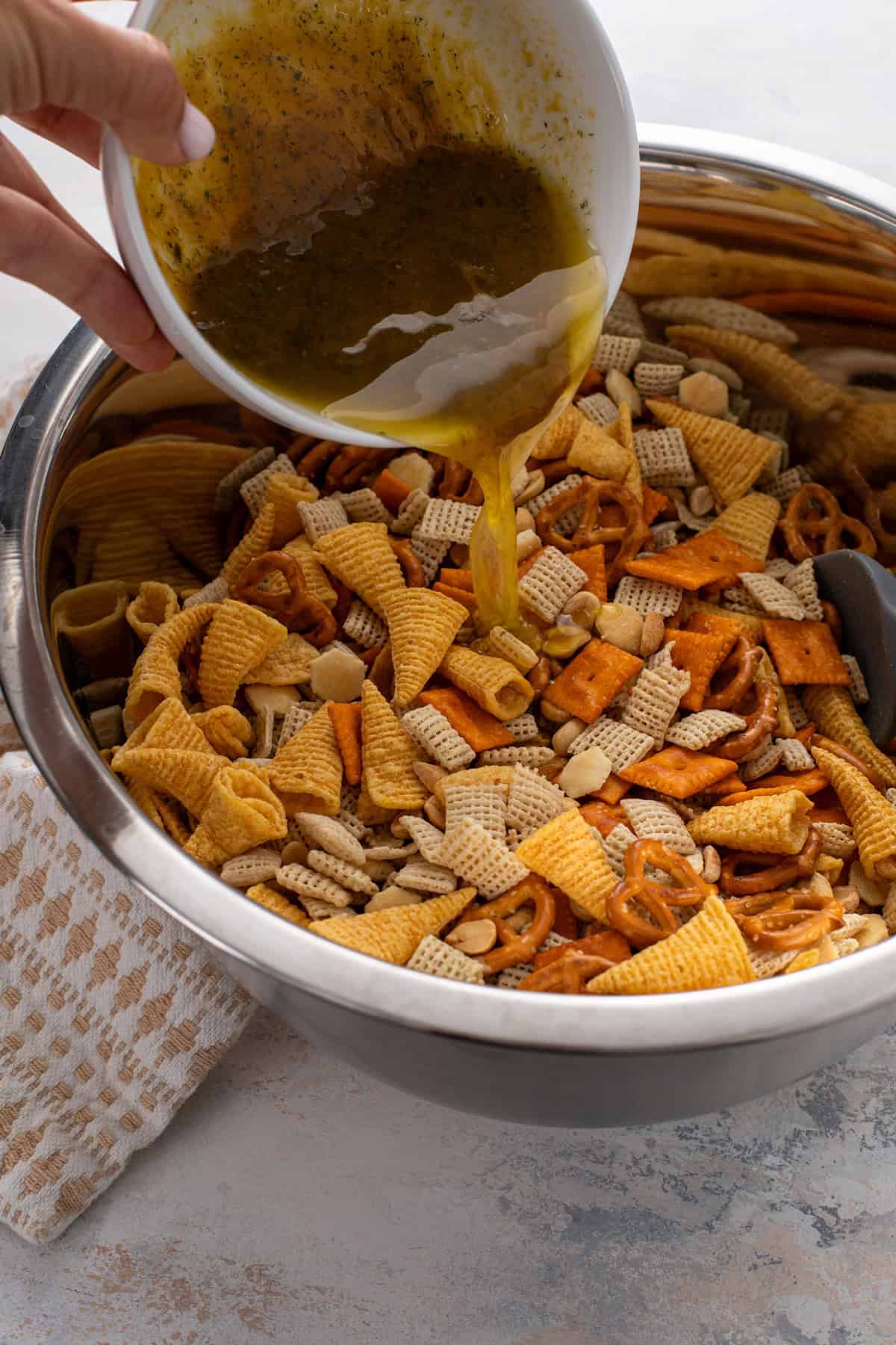 Ranch chex mix seasonings being drizzled over the cereal mixture in a large bowl.