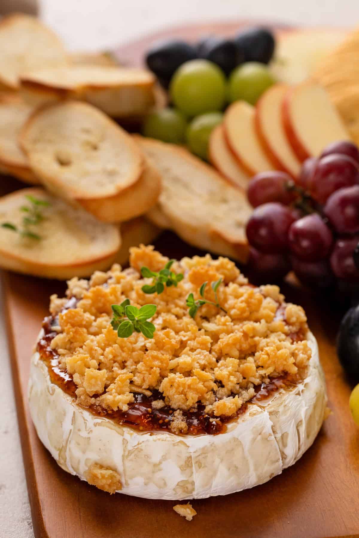 Wheel of baked brie with jam on a wooden board with fruit and baguette slices in the background.